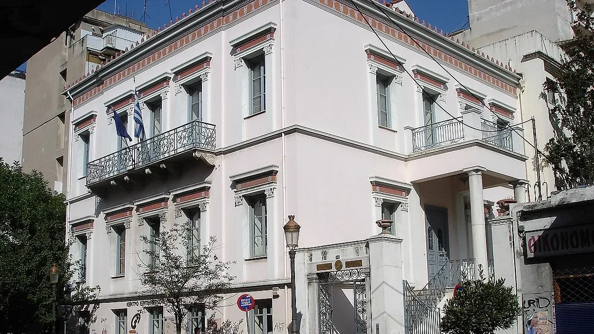 Νεοκλασικό διώροφο κτίριο με λευκή πρόσοψη, μπαλκόνια με μεταλλικά κάγκελα και ελληνικές σημαίες.