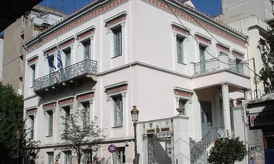 Νεοκλασικό διώροφο κτίριο με λευκή πρόσοψη, μπαλκόνια με μεταλλικά κάγκελα και ελληνικές σημαίες.