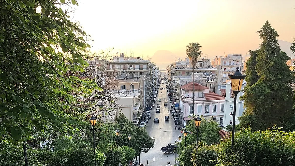 Αστικό τοπίο με κτίρια, δρόμο με αυτοκίνητα και δέντρα υπό το φως του ηλιοβασιλέματος.