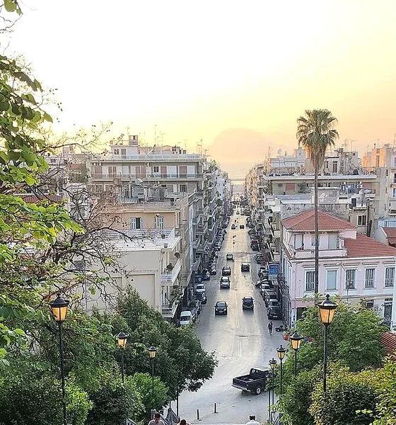 Αστικό τοπίο με κτίρια, δρόμο με αυτοκίνητα και δέντρα υπό το φως του ηλιοβασιλέματος.