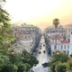 Αστικό τοπίο με κτίρια, δρόμο με αυτοκίνητα και δέντρα υπό το φως του ηλιοβασιλέματος.