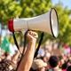 Χέρι κρατά λευκό και κόκκινο μεγάφωνο μπροστά από συγκεντρωμένο πλήθος με σημαίες
