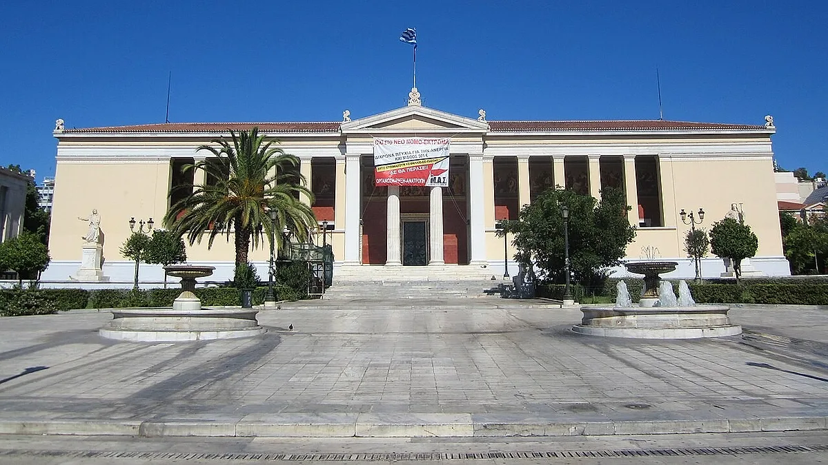 Το κτήριο του Πανεπιστημίου Αθηνών με πανό στην πρόσοψη, μπροστά από πλακόστρωτη πλατεία με σιντριβάνια και φοίνικα