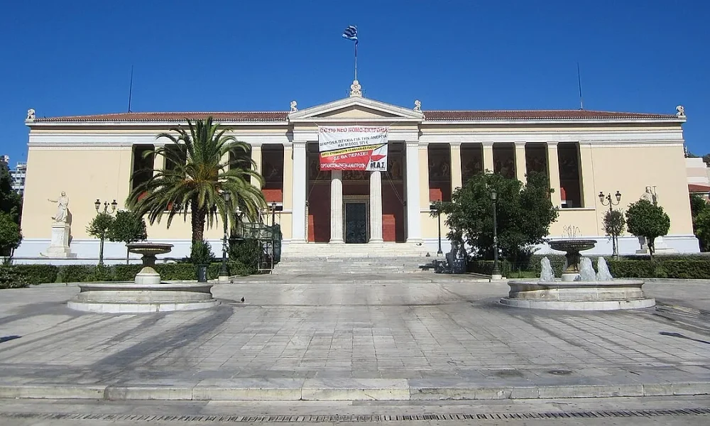 Το κτήριο του Πανεπιστημίου Αθηνών με πανό στην πρόσοψη, μπροστά από πλακόστρωτη πλατεία με σιντριβάνια και φοίνικα