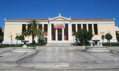 Το κτήριο του Πανεπιστημίου Αθηνών με πανό στην πρόσοψη, μπροστά από πλακόστρωτη πλατεία με σιντριβάνια και φοίνικα