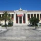 Το κτήριο του Πανεπιστημίου Αθηνών με πανό στην πρόσοψη, μπροστά από πλακόστρωτη πλατεία με σιντριβάνια και φοίνικα