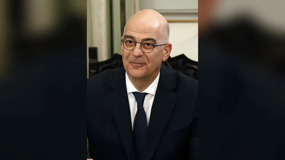Άντρας με γυαλιά, σκούρο κοστούμι, λευκό πουκάμισο και μπλε γραβάτα ποζάρει μπροστά σε εσωτερικό χώρο.