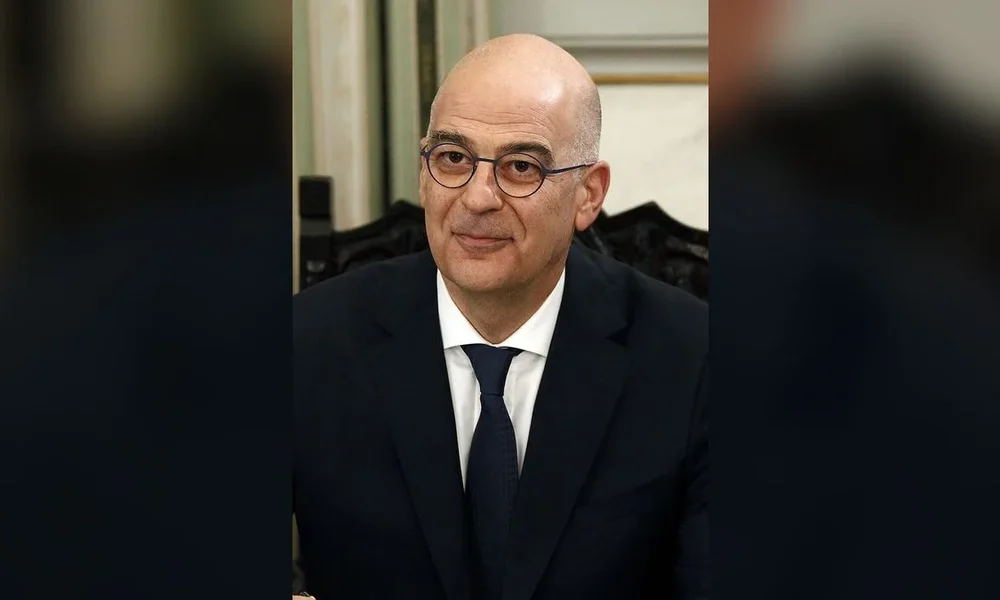 Άντρας με γυαλιά, σκούρο κοστούμι, λευκό πουκάμισο και μπλε γραβάτα ποζάρει μπροστά σε εσωτερικό χώρο.
