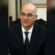Άντρας με γυαλιά, σκούρο κοστούμι, λευκό πουκάμισο και μπλε γραβάτα ποζάρει μπροστά σε εσωτερικό χώρο.