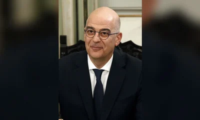 Άντρας με γυαλιά, σκούρο κοστούμι, λευκό πουκάμισο και μπλε γραβάτα ποζάρει μπροστά σε εσωτερικό χώρο.