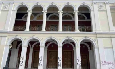 Η πρόσοψη κτιρίου με κολόνες, καμάρες, μπαλκόνι και ξύλινες πόρτες, όπου διακρίνονται συνθήματα σε λευκές κολόνες.