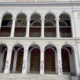 Η πρόσοψη κτιρίου με κολόνες, καμάρες, μπαλκόνι και ξύλινες πόρτες, όπου διακρίνονται συνθήματα σε λευκές κολόνες.