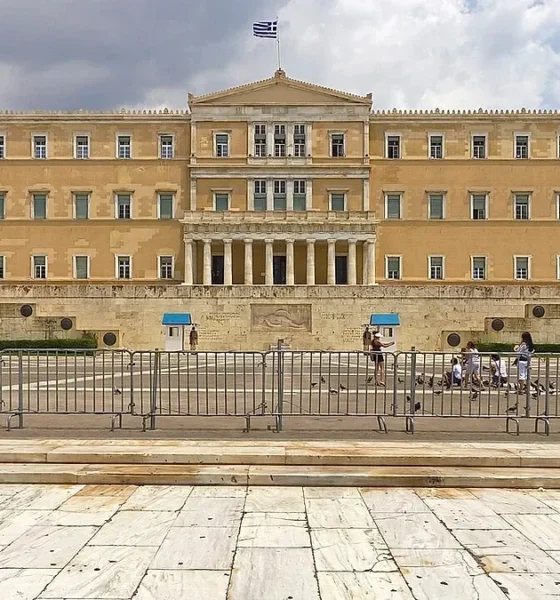 Το κτίριο της Βουλής των Ελλήνων στην Αθήνα, μπροστά από το οποίοι υπάρχουν προστατευτικά κιγκλιδώματα και περαστικοί.