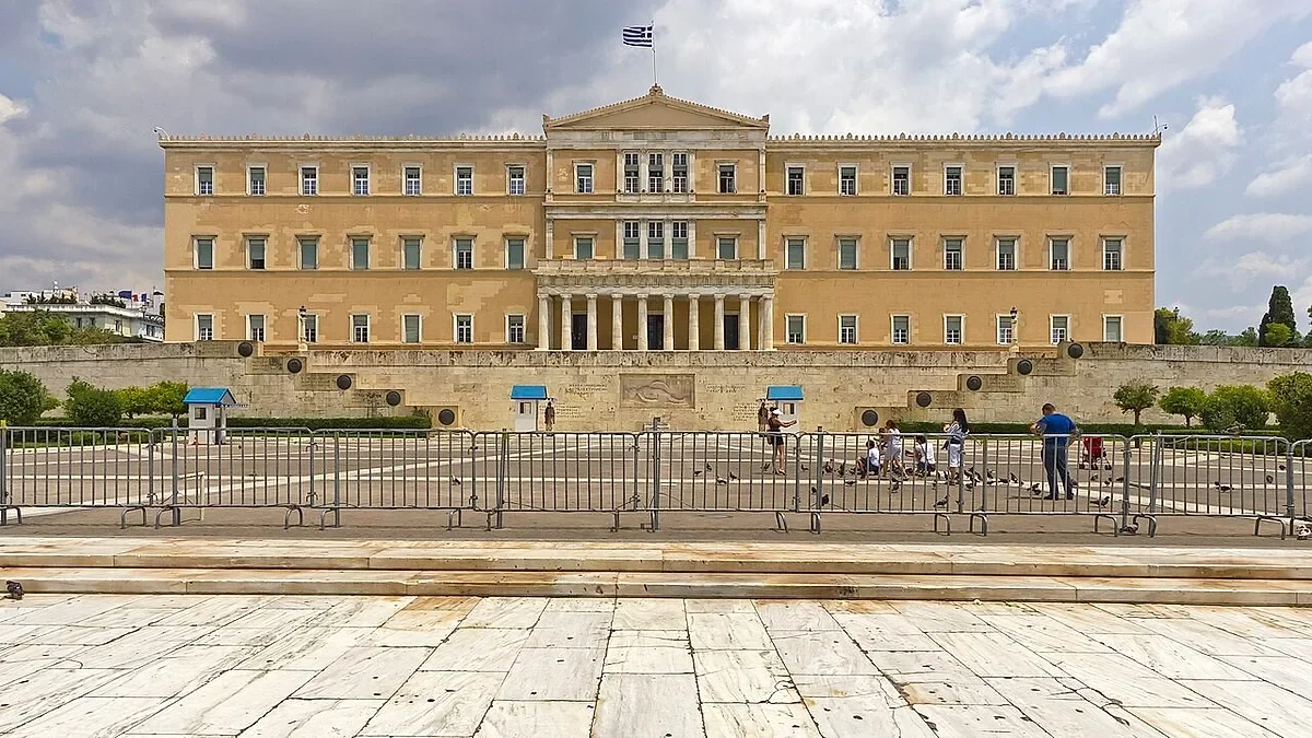 Το κτίριο της Βουλής των Ελλήνων στην Αθήνα, μπροστά από το οποίοι υπάρχουν προστατευτικά κιγκλιδώματα και περαστικοί.