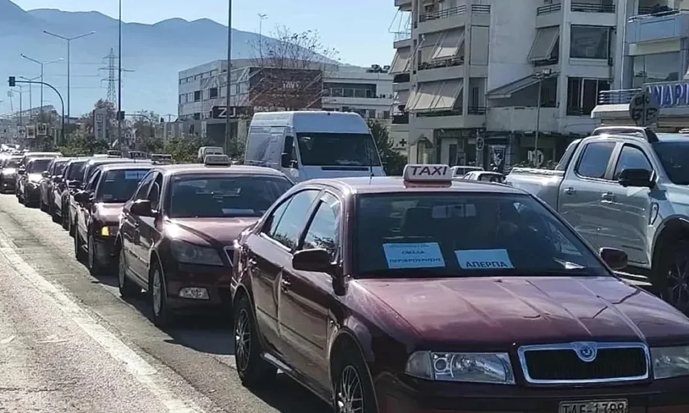 Σειρά από σταθμευμένα αυτοκίνητα ταξί σε δρόμο πόλης με κίνηση και κτίρια στο βάθος.