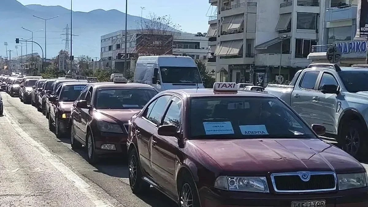 Σειρά από σταθμευμένα αυτοκίνητα ταξί σε δρόμο πόλης με κίνηση και κτίρια στο βάθος.