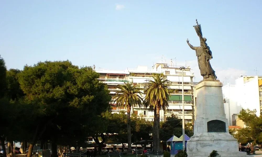 Άποψη πλατείας με ψηλό άγαλμα σε βάθρο, φοίνικες, δέντρα και πολυκατοικίες στο βάθος κάτω από ανοιχτόχρωμο ουρανό.