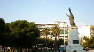 Άποψη πλατείας με ψηλό άγαλμα σε βάθρο, φοίνικες, δέντρα και πολυκατοικίες στο βάθος κάτω από ανοιχτόχρωμο ουρανό.