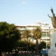 Άποψη πλατείας με ψηλό άγαλμα σε βάθρο, φοίνικες, δέντρα και πολυκατοικίες στο βάθος κάτω από ανοιχτόχρωμο ουρανό.