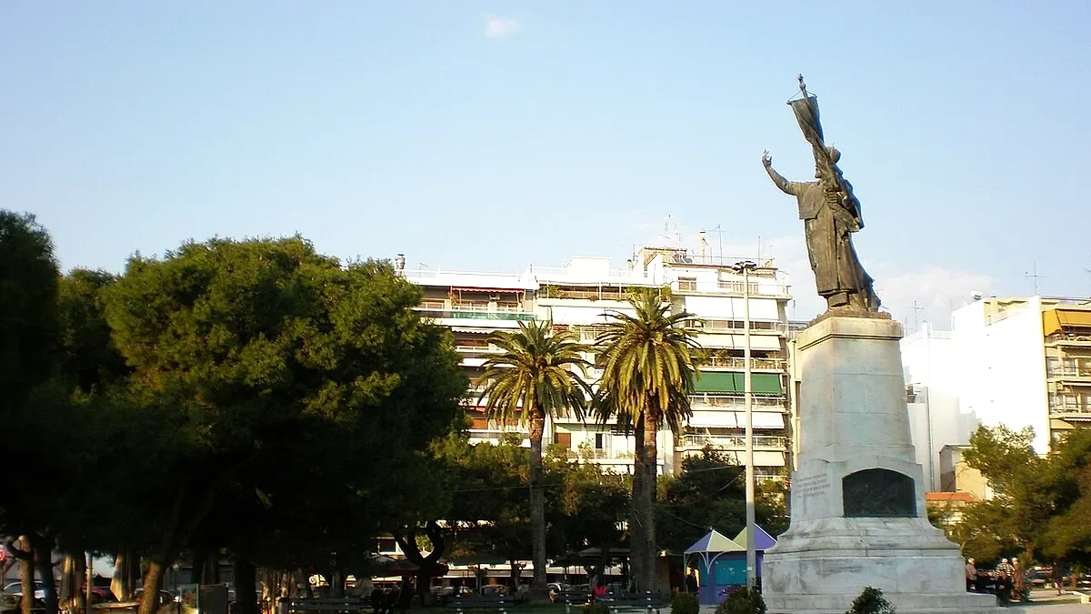 Άποψη πλατείας με ψηλό άγαλμα σε βάθρο, φοίνικες, δέντρα και πολυκατοικίες στο βάθος κάτω από ανοιχτόχρωμο ουρανό.