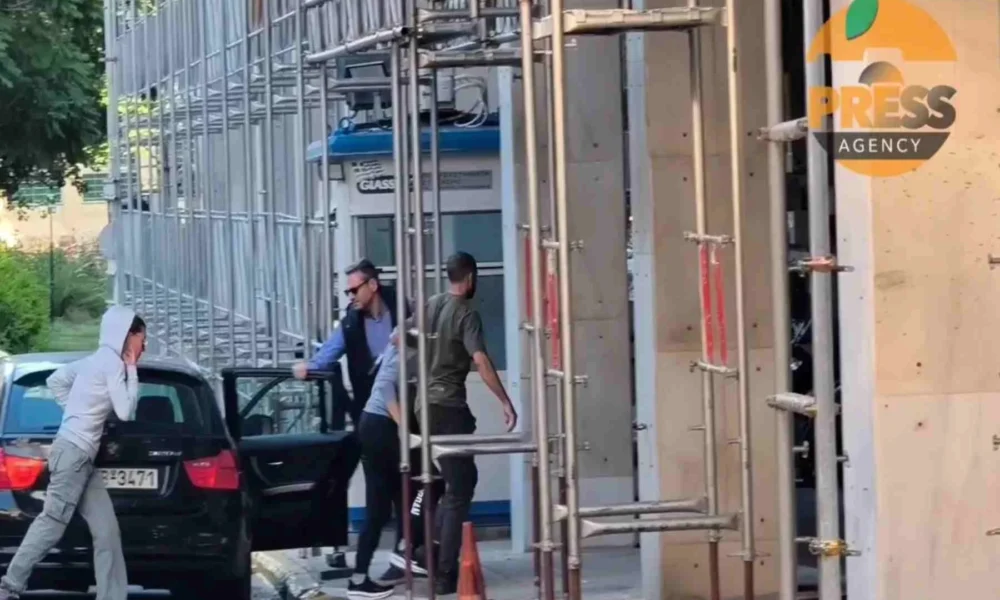 Άτομα κινούνται κοντά σε αυτοκίνητο και οικοδομική σκαλωσιά δίπλα σε κτήριο με φυλάκιο.