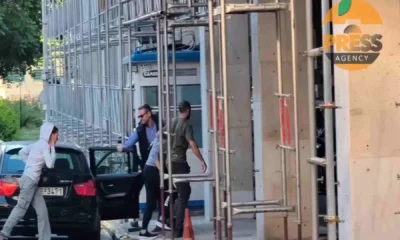 Άτομα κινούνται κοντά σε αυτοκίνητο και οικοδομική σκαλωσιά δίπλα σε κτήριο με φυλάκιο.