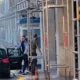Άτομα κινούνται κοντά σε αυτοκίνητο και οικοδομική σκαλωσιά δίπλα σε κτήριο με φυλάκιο.