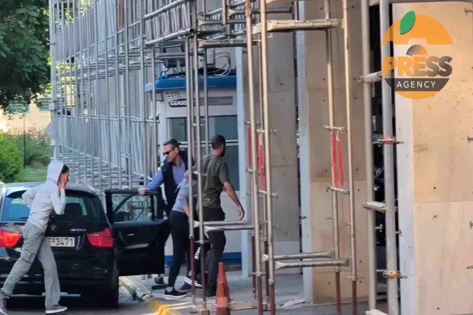 Άτομα κινούνται κοντά σε αυτοκίνητο και οικοδομική σκαλωσιά δίπλα σε κτήριο με φυλάκιο.