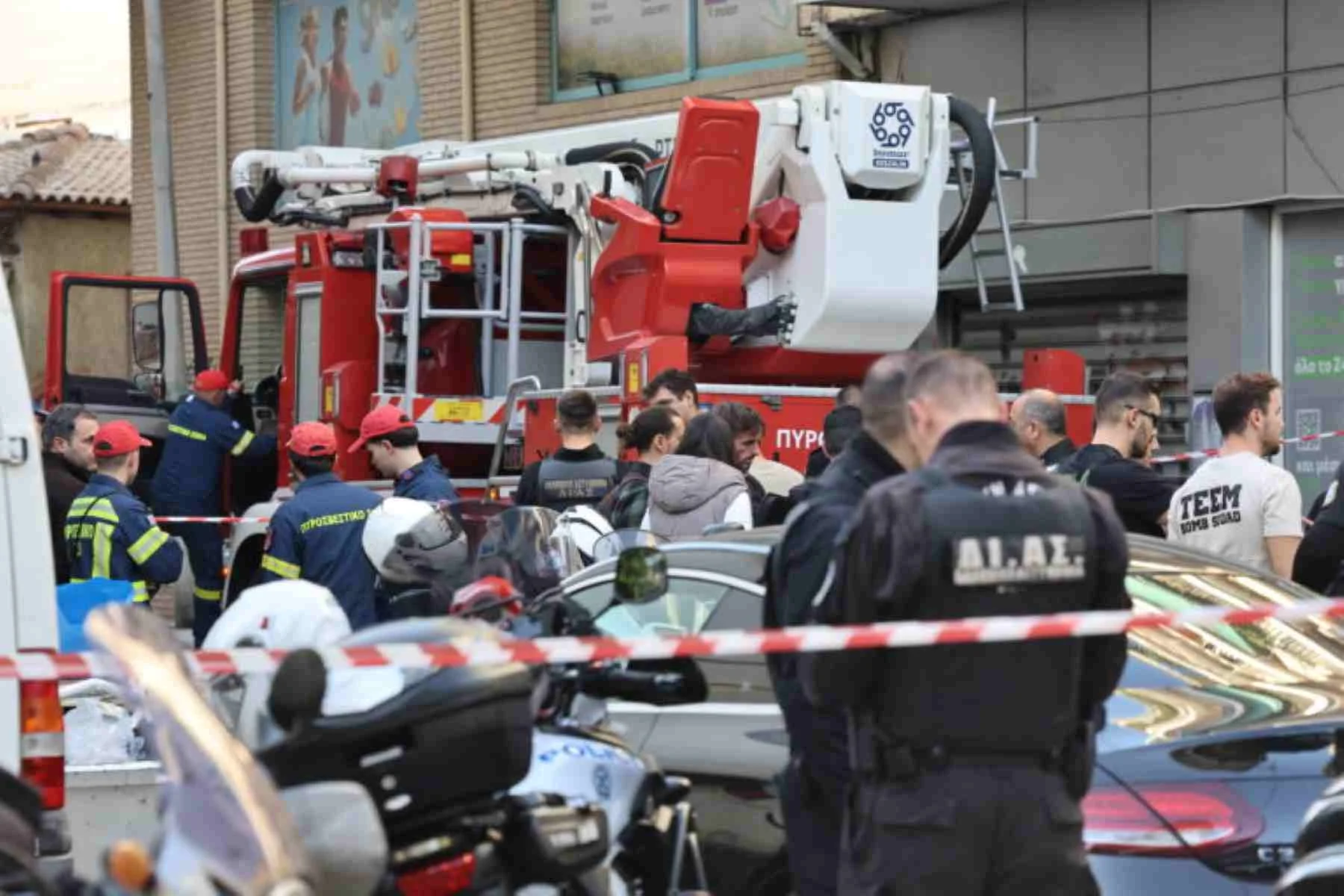 Πλήθος κόσμου και αστυνομικοί συγκεντρωμένοι σε δρόμο μπροστά από όχημα της πυροσβεστικής και αυτοκίνητα, αποκλεισμένοι με κόκκινη