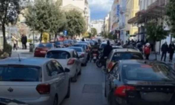 Αυτοκίνητα και δίκυκλα ακινητοποιημένα σε δρόμο πόλης με κίνηση και πεζούς στα πεζοδρόμια.