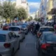 Αυτοκίνητα και δίκυκλα ακινητοποιημένα σε δρόμο πόλης με κίνηση και πεζούς στα πεζοδρόμια.