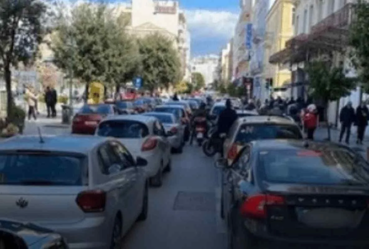 Αυτοκίνητα και δίκυκλα ακινητοποιημένα σε δρόμο πόλης με κίνηση και πεζούς στα πεζοδρόμια.