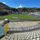 Πανοραμική άποψη υπαίθριου γηπέδου ποδοσφαίρου με κερκίδες και λόφους στο βάθος υπό ηλιοφάνεια.