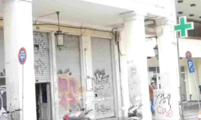 Πρόσοψη κτιρίου με κλειστά ρολά, κολόνες, γκράφιτι, πινακίδα φαρμακείου και σταθμευμένα δίκυκλα στο πεζοδρόμιο.