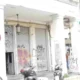 Πρόσοψη κτιρίου με κλειστά ρολά, κολόνες, γκράφιτι, πινακίδα φαρμακείου και σταθμευμένα δίκυκλα στο πεζοδρόμιο.