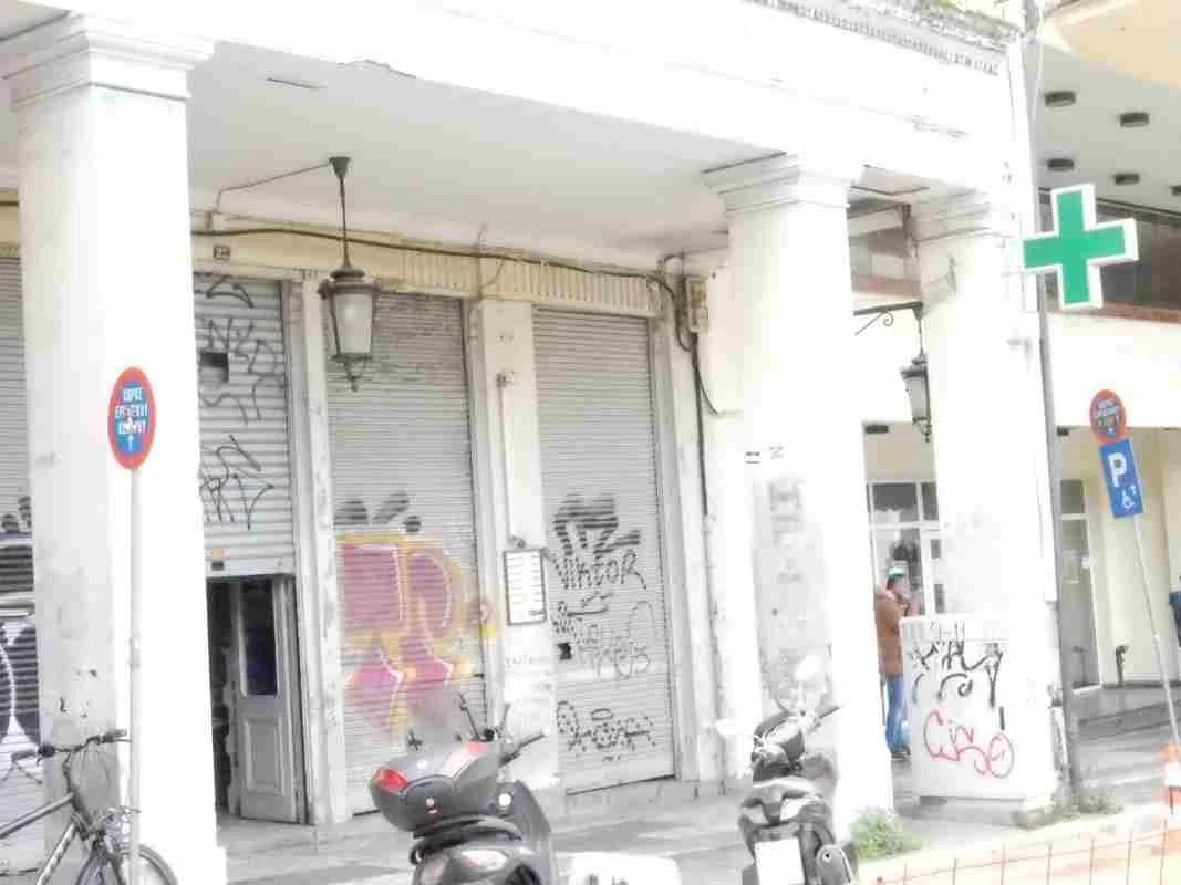 Πρόσοψη κτιρίου με κλειστά ρολά, κολόνες, γκράφιτι, πινακίδα φαρμακείου και σταθμευμένα δίκυκλα στο πεζοδρόμιο.