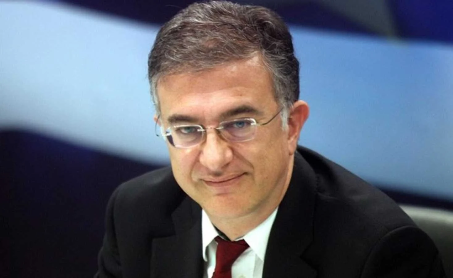 Άντρας με γυαλιά οράσεως, κοστούμι και κόκκινη γραβάτα κοιτάζει μπροστά.