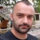 Πορτραίτο άνδρα με κοντά μαλλιά και μούσι, ο οποίος φοράει σκούρο μπλουζάκι σε εξωτερικό χώρο.