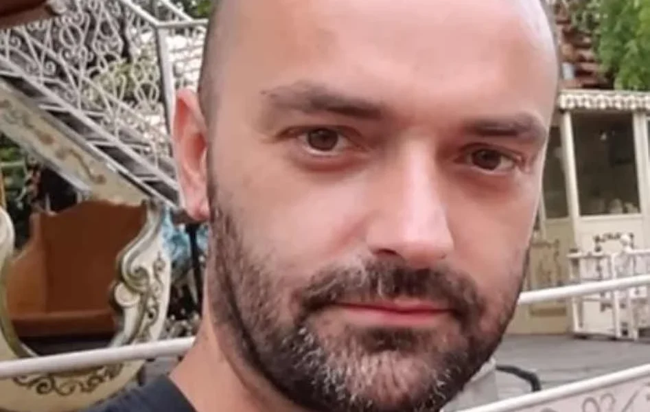 Πορτραίτο άνδρα με κοντά μαλλιά και μούσι, ο οποίος φοράει σκούρο μπλουζάκι σε εξωτερικό χώρο.