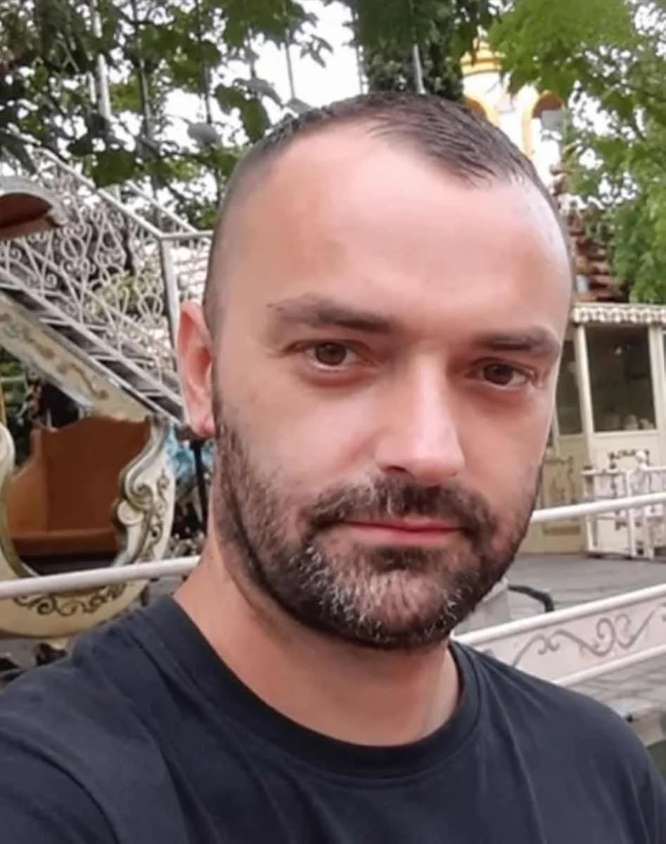 Πορτραίτο άνδρα με κοντά μαλλιά και μούσι, ο οποίος φοράει σκούρο μπλουζάκι σε εξωτερικό χώρο.
