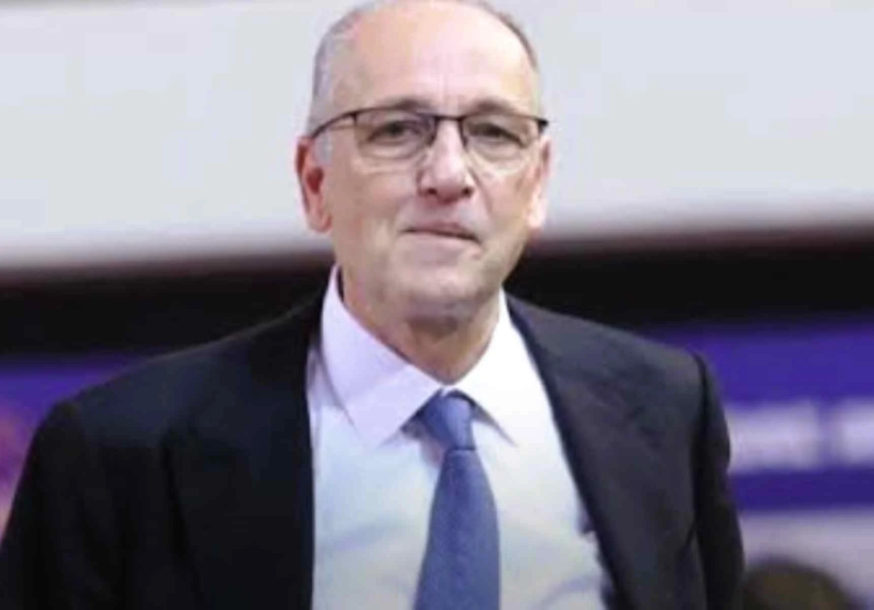 Άντρας με γυαλιά, λευκό πουκάμισο, μπλε γραβάτα και σκούρο σακάκι κοιτάζει μπροστά.