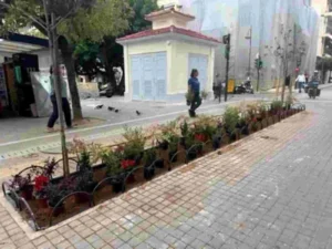 Πεζόδρομος με πλακόστρωτο, παρτέρια με φυτά, μικρό κτίριο στο βάθος και περαστικοί.