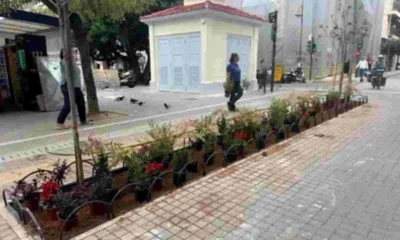 Πεζόδρομος με πλακόστρωτο, παρτέρια με φυτά, μικρό κτίριο στο βάθος και περαστικοί.