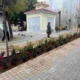Πεζόδρομος με πλακόστρωτο, παρτέρια με φυτά, μικρό κτίριο στο βάθος και περαστικοί.