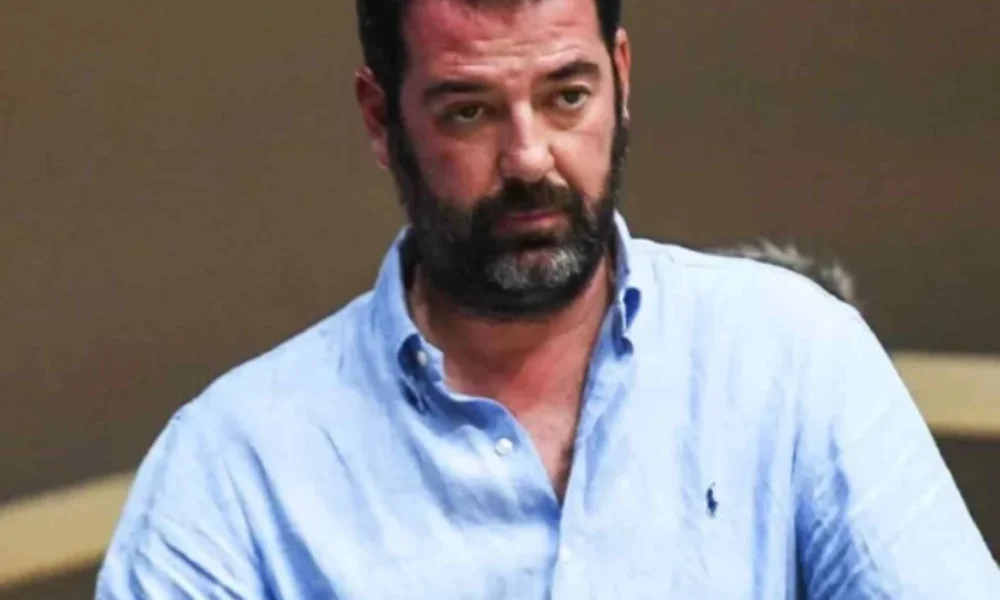 Άντρας με γένια και γαλάζιο πουκάμισο κοιτάζει μπροστά.