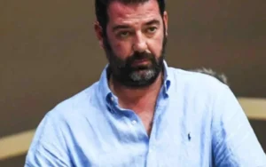 Άντρας με γένια και γαλάζιο πουκάμισο κοιτάζει μπροστά.