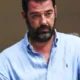 Άντρας με γένια και γαλάζιο πουκάμισο κοιτάζει μπροστά.
