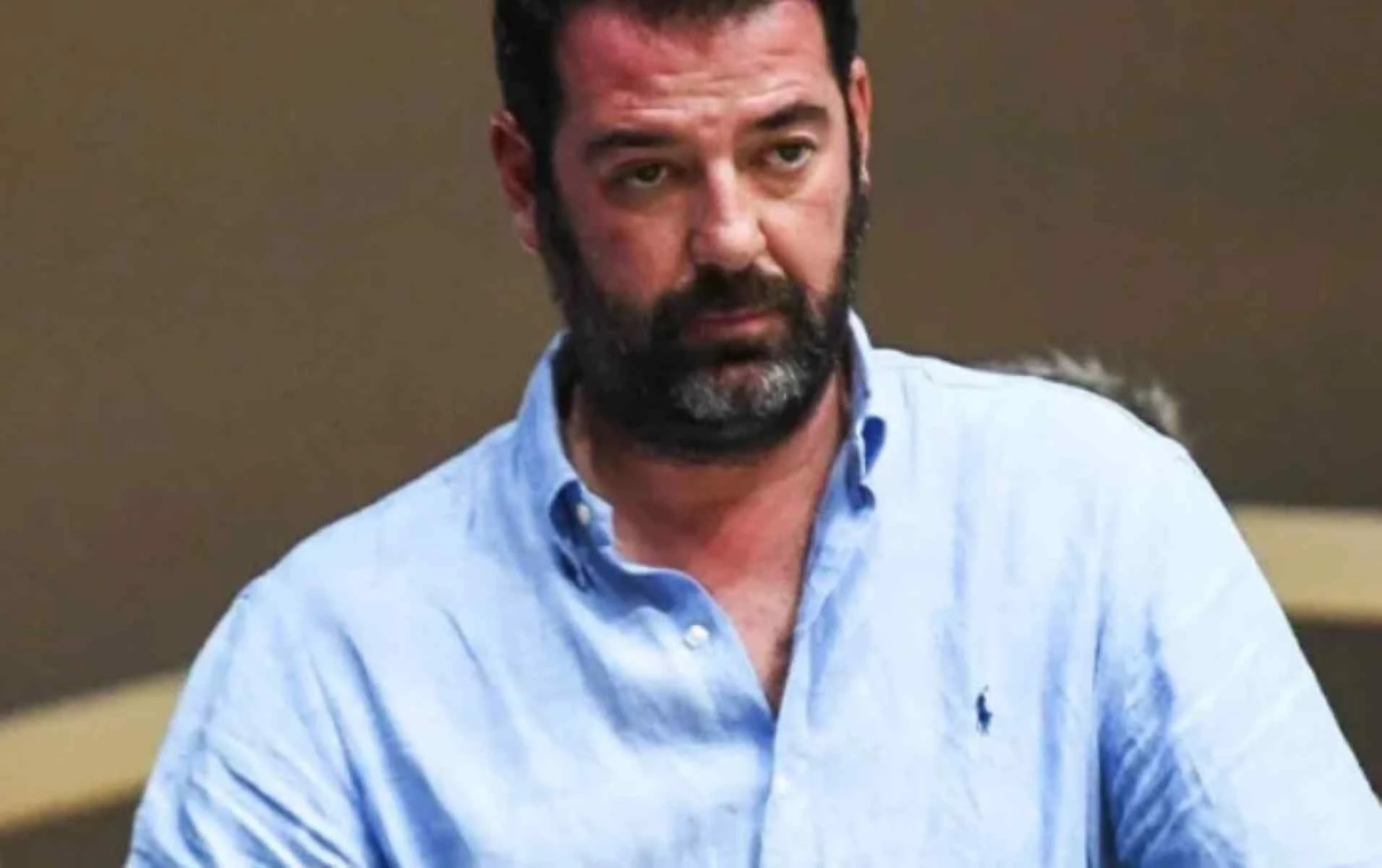 Άντρας με γένια και γαλάζιο πουκάμισο κοιτάζει μπροστά.