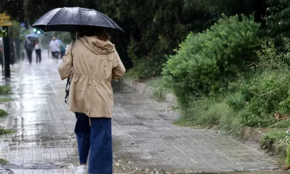 Άτομο με ομπρέλα και αδιάβροχο περπατά σε βρεγμένο πεζοδρόμιο υπό βροχή