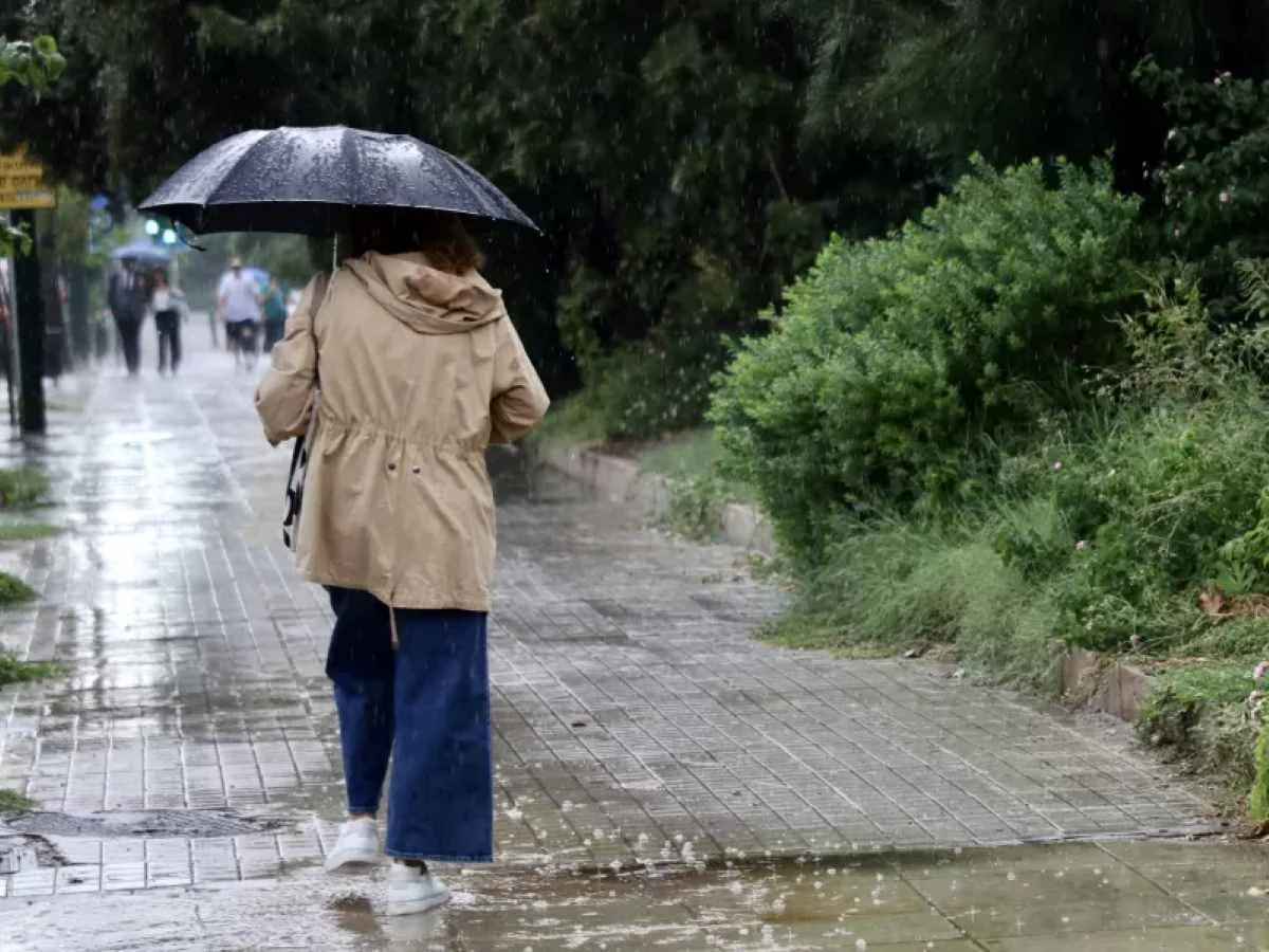 Άτομο με ομπρέλα και αδιάβροχο περπατά σε βρεγμένο πεζοδρόμιο υπό βροχή